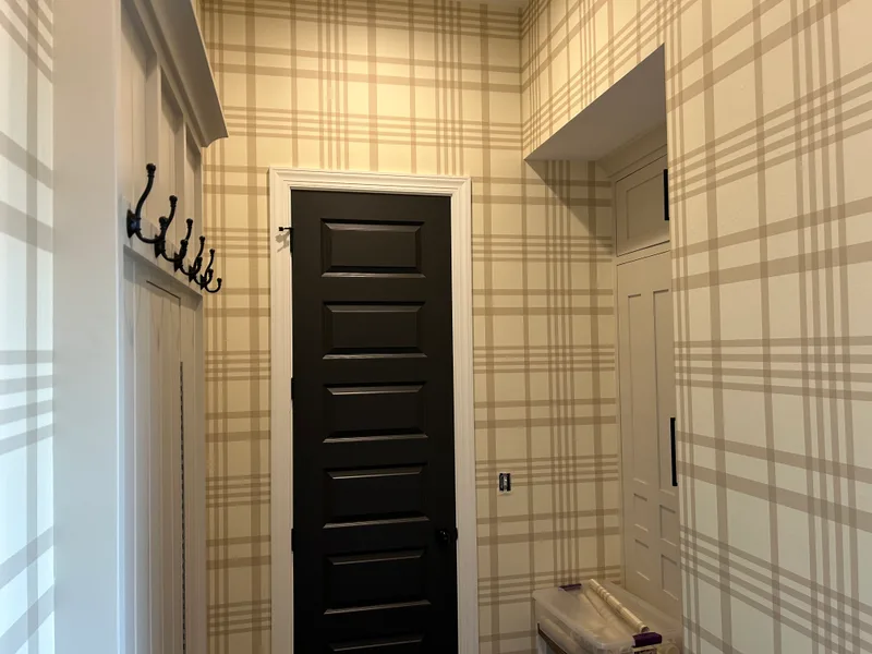 This mudroom features a classic plaid wallpaper in soft beige and cream tones, creating a warm, welcoming, and timeless atmosphere. The large-scale grid pattern adds subtle structure and visual interest while maintaining a light and airy feel that complements the white trim, dark door, and built-in cabinetry.