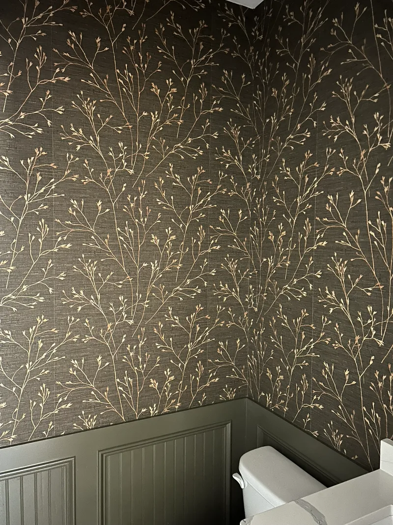 This sophisticated powder room features a luxurious printed faux grasscloth wallpaper with a delicate, branching botanical motif in metallic gold on a deep charcoal background. The elegant pattern adds subtle shimmer and texture, beautifully contrasting with the dark wainscoting below for a dramatic and refined look.