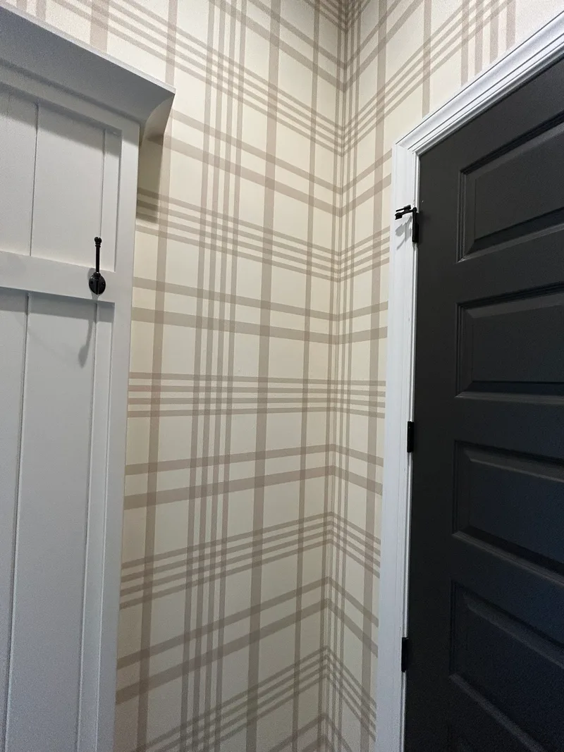 This mudroom features a classic plaid wallpaper in soft beige and cream tones, creating a warm, welcoming, and timeless atmosphere. The large-scale grid pattern adds subtle structure and visual interest while maintaining a light and airy feel that complements the white trim, dark door, and built-in cabinetry.