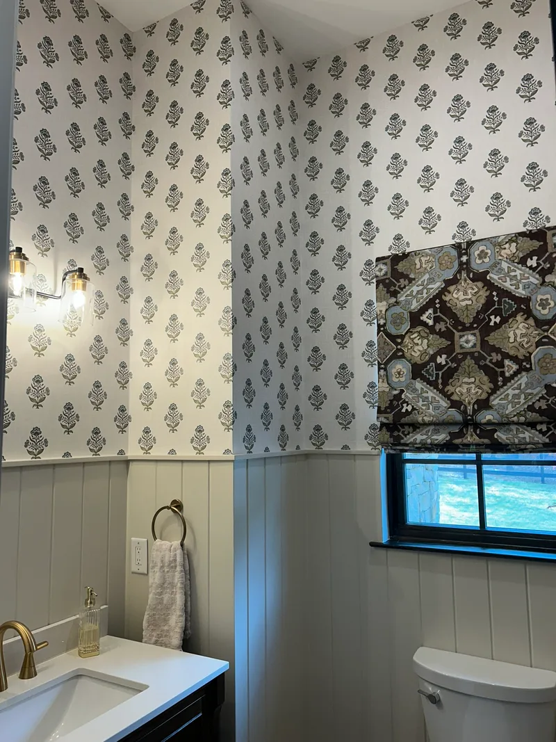 This elegant powder room features a sophisticated botanical wallpaper with a delicate repeating floral motif in soft neutral tones. The wallpaper covers the upper walls, creating a timeless and refined backdrop that pairs beautifully with the crisp white wainscoting below. A bold patterned Roman shade complements the wallpaper's design, adding depth and visual interest to the space.
