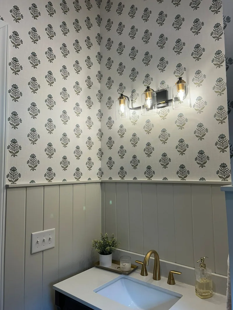This elegant powder room features a sophisticated botanical wallpaper with a delicate repeating floral motif in soft neutral tones. The wallpaper covers the upper walls, creating a timeless and refined backdrop that pairs beautifully with the crisp white wainscoting below. A bold patterned Roman shade complements the wallpaper's design, adding depth and visual interest to the space.